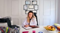 Nutritionist sits at a modern office desk writing, with fruits and diplomas displayed, emphasizing health and professionalism.