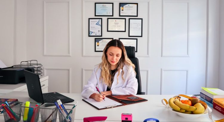 Nutritionist sits at a modern office desk writing, with fruits and diplomas displayed, emphasizing health and professionalism.